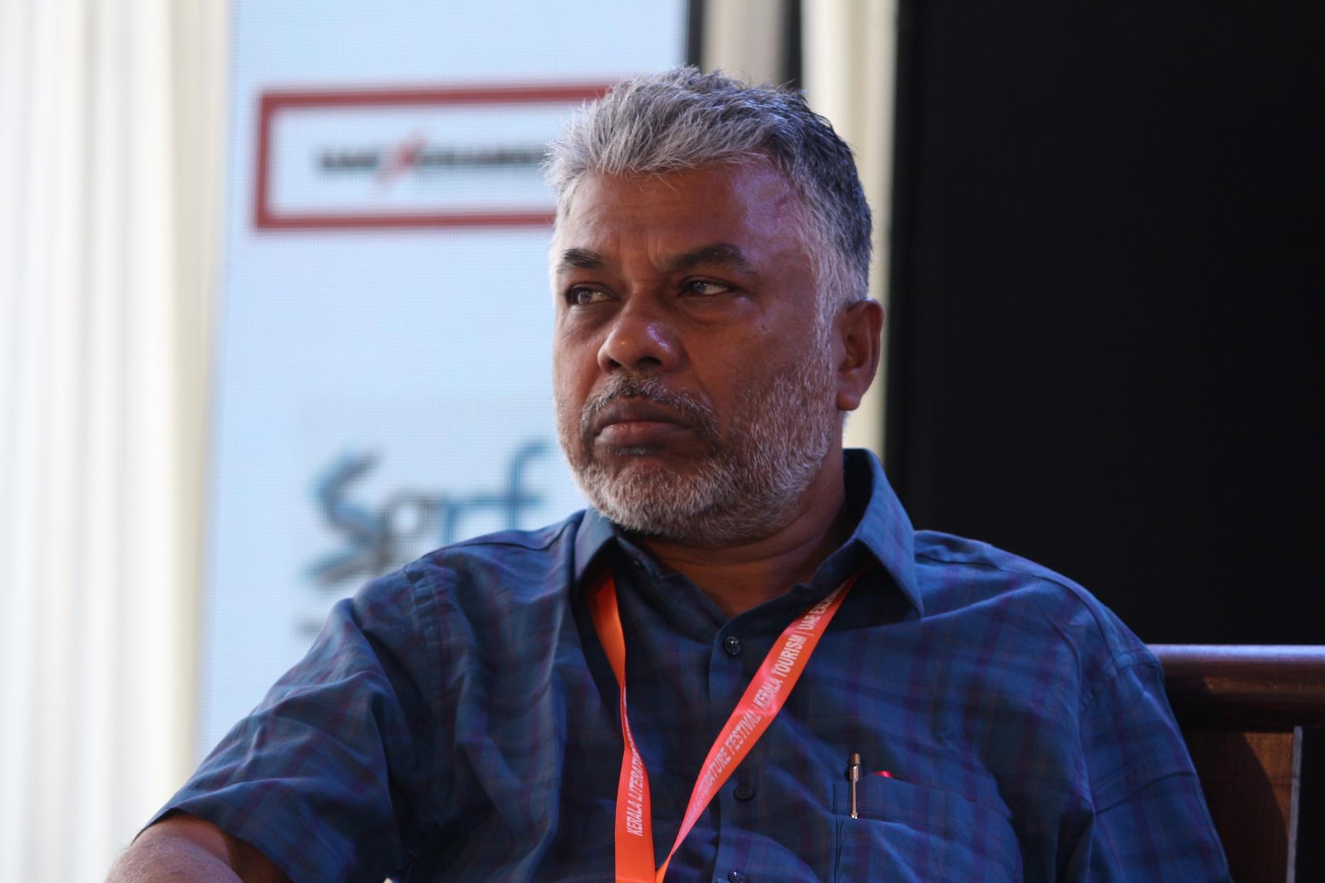 Perumalmurugan seated on stage in a blue checked shirt, looking to one side